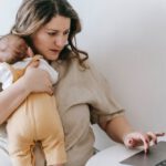 Home Business - Concentrated young female freelancer embracing newborn while sitting at table and working remotely on laptop at home