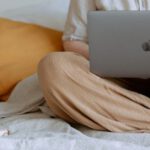 Home Business - Crop faceless female in casual outfit sitting on bed with legs crossed holding laptop on knees with cup of coffee standing on notebook while working from home