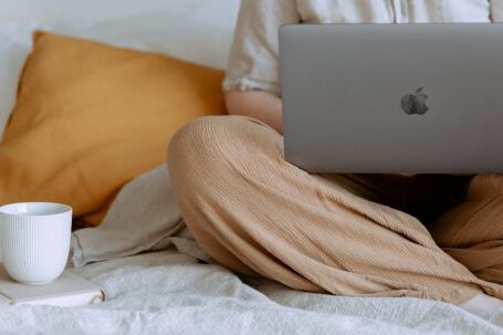 Home Business - Crop faceless female in casual outfit sitting on bed with legs crossed holding laptop on knees with cup of coffee standing on notebook while working from home