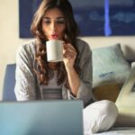 Home Business - Woman in Grey Jacket Sits on Bed Uses Grey Laptop