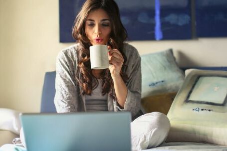 Home Business - Woman in Grey Jacket Sits on Bed Uses Grey Laptop