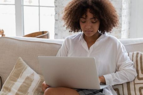 Home Business - Full body of young stylish barefooted black female freelancer with Afro hair in casual outfit working remotely on laptop while sitting on comfortable sofa in cozy living room