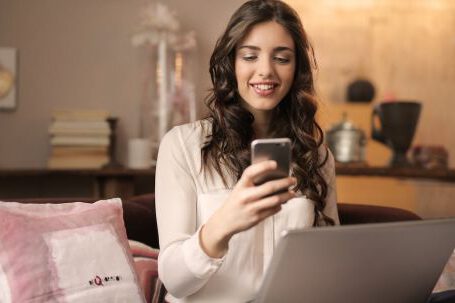 Home Business - Woman Sitting on Sofa While Looking at Phone With Laptop on Lap