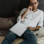 Home Office - A Man in White Long Sleeves and Denim Jeans Resting on the Bed while Having a Phone Call