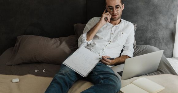 Home Office - A Man in White Long Sleeves and Denim Jeans Resting on the Bed while Having a Phone Call
