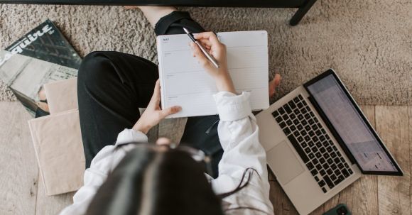 Home Business - Top view of anonymous woman in casual wear sitting on floor with laptop and smartphone and creating plan on notebook while resting during break in modern living room